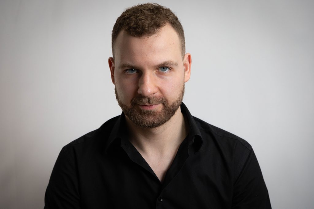 Portret pianisty w czarnej koszuli, z krótkimi włosami i brodą, wykonany w studio na jasnym tle.