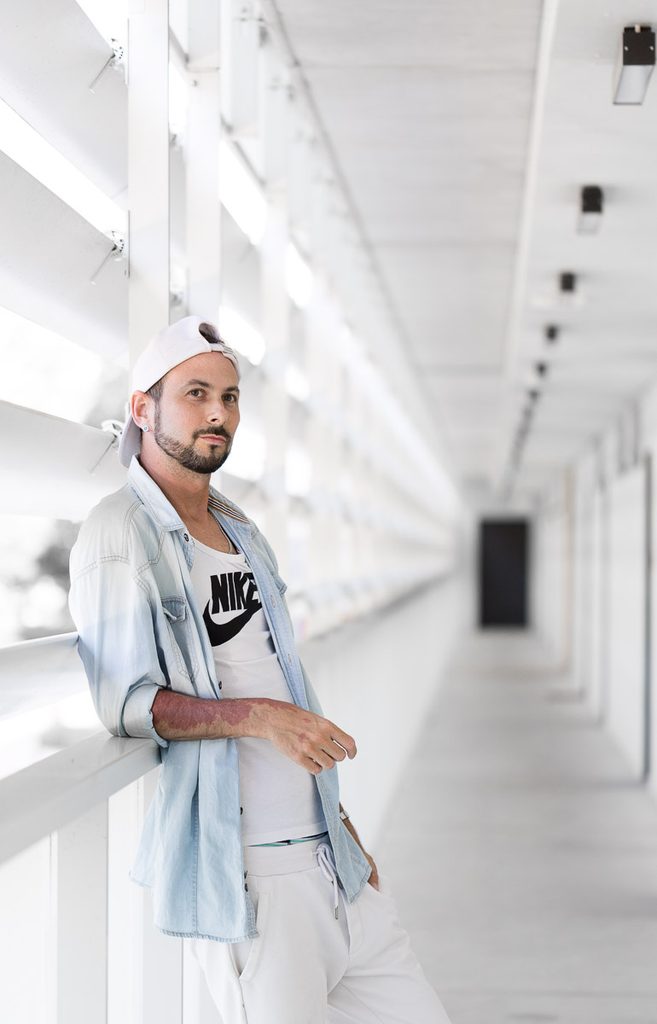 Portret fotografa ze Szczecina w jasnym wnętrzu, ubranego w białą koszulkę, rozpiętą koszulę i czapkę z daszkiem.