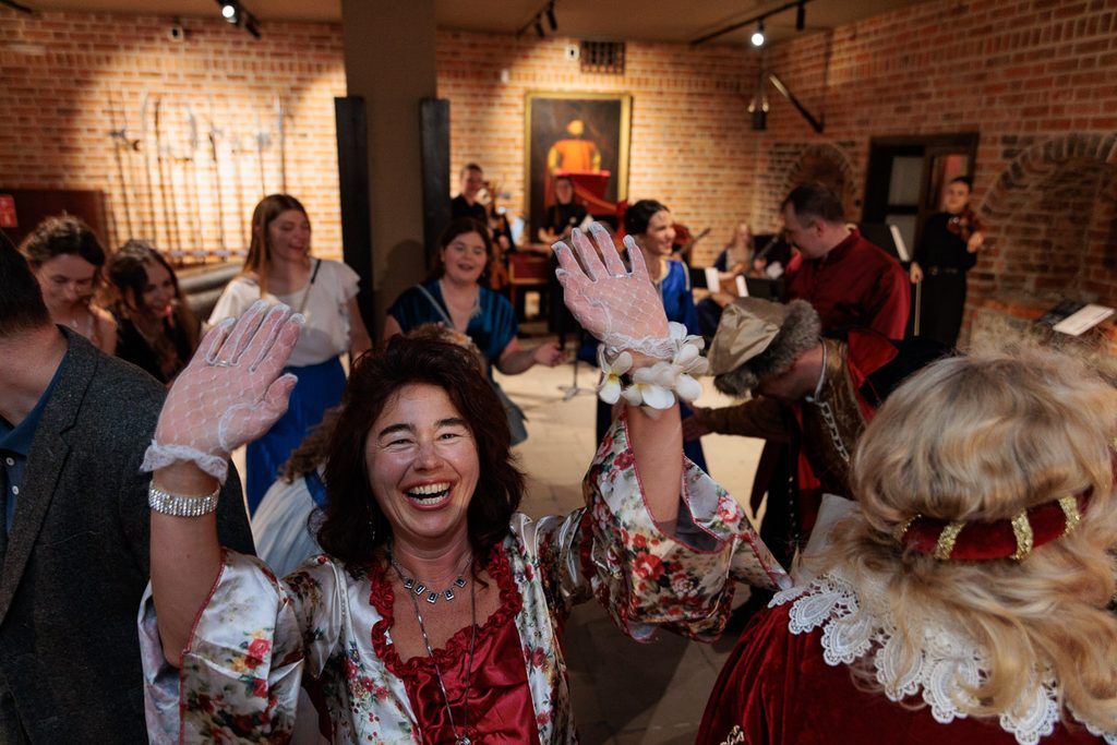 Uśmiechnięta uczestniczka festiwalu strojów dawnych z uniesionymi rękami podczas wydarzenia w historycznym wnętrzu.