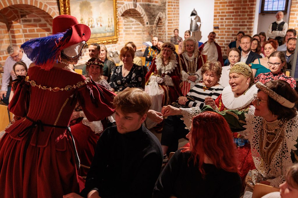 Uczestnicy balu w historycznych kostiumach podczas barwnego wydarzenia artystycznego w zabytkowym ceglanym wnętrzu.
