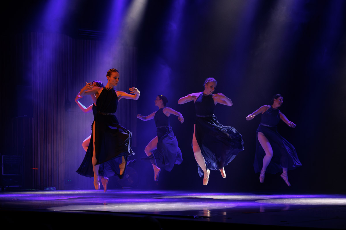 Zespół tancerzy wykonujących dynamiczną choreografię na scenie w niebieskim świetle reflektorów.