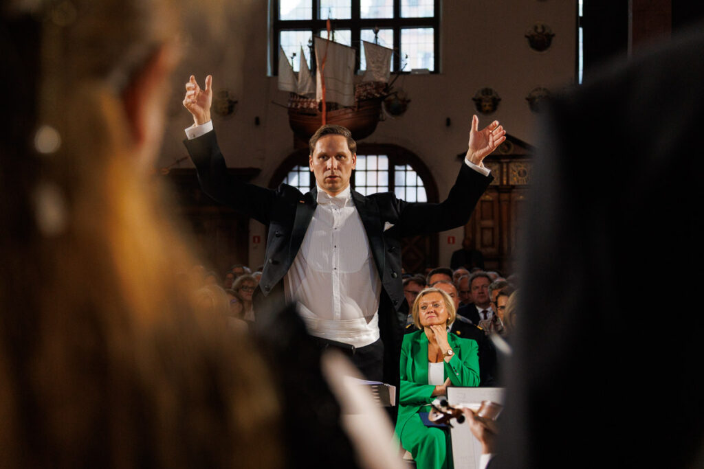 Dyrygent Rafał Kłoczko podczas prowadzenia koncertu wśród publiczności – fotografia dyrygenta i wydarzenia muzycznego.