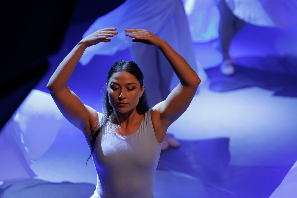 Tancerka w białym kostiumie wykonująca choreografię z uniesionymi ramionami w niebieskim świetle scenicznym.