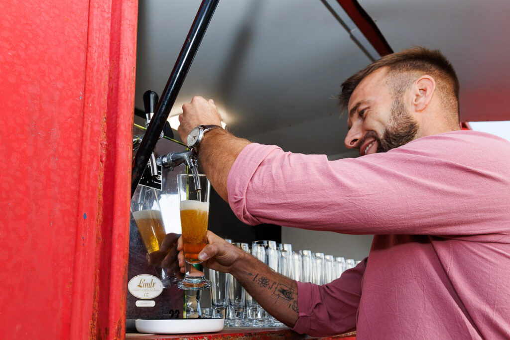 Barman w różowej koszuli nalewający piwo z kranu podczas obsługi baru na wydarzeniu plenerowym.